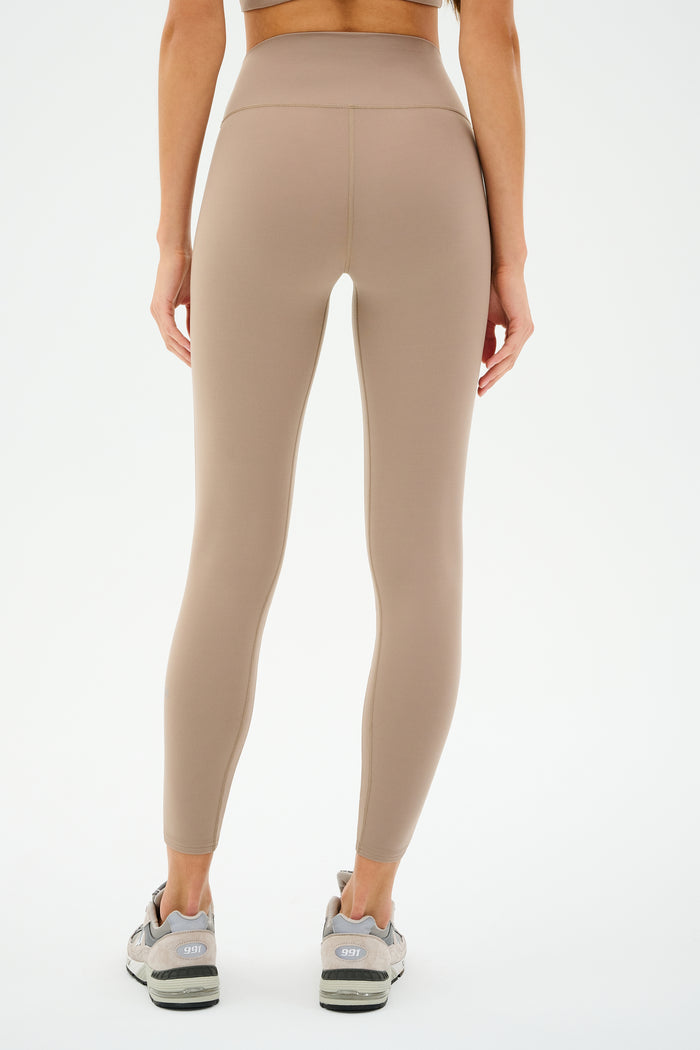 A person stands with their back to the camera against a white backdrop, wearing SPLITS59 Chevron Airweight High Waist 7/8 leggings in Latte/Indigo and gray athletic shoes.