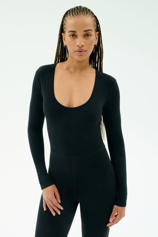 A woman with long braided hair wears the SPLITS59 Chloe Airweight Jumpsuit in black, standing against a plain light background.