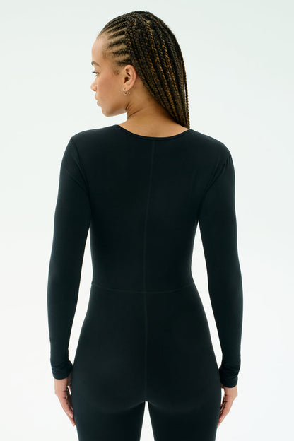 A person with braided hair wears the SPLITS59 Chloe Airweight Jumpsuit in black, made from soft Airweight fabric, standing against a plain light background.