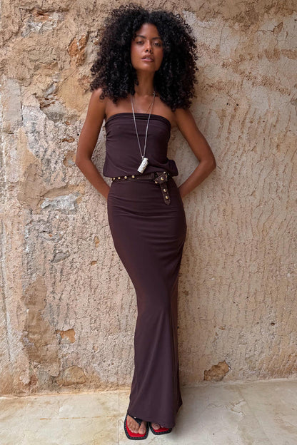 Chocolate Sabine Dress in brown, styled as a strapless column maxi dress on a model against a rustic wall.