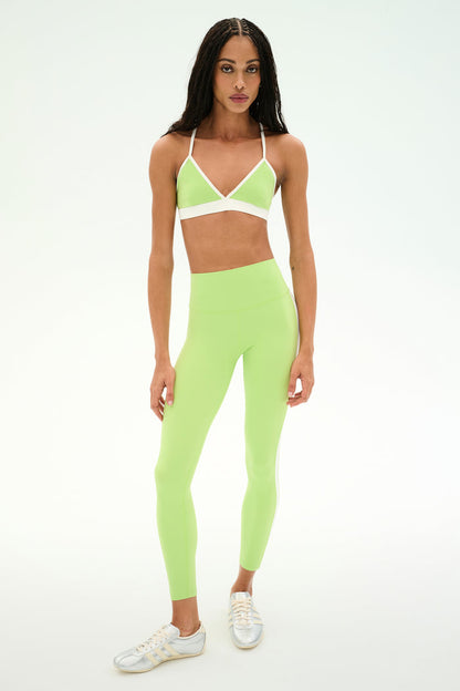 A woman stands facing forward against a plain light background wearing the SPLITS59 Clare High Waist Rigor 7/8 leggings and matching sports bra set in Limeade/White, crafted from a soft nylon-spandex blend, and paired with white sneakers.