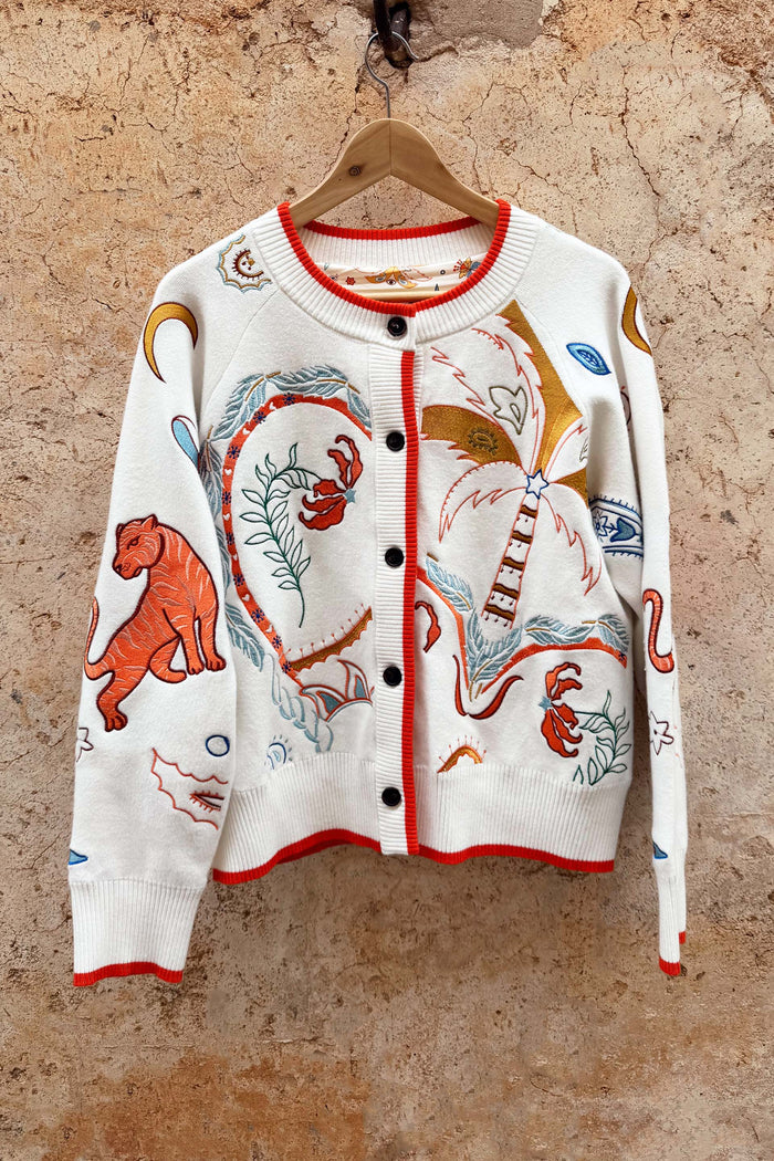 Cream knit Cheryl Cardigan with orange contrast trims and embroidered tigress and palm motifs on a wooden hanger.
