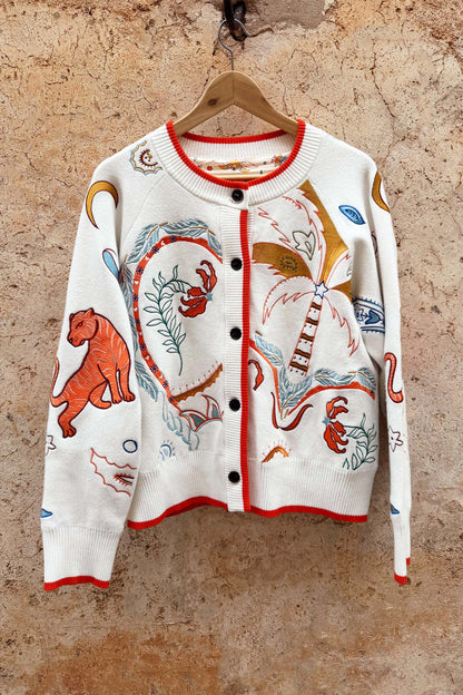 Cream knit Cheryl Cardigan with orange contrast trims and embroidered tigress and palm motifs on a wooden hanger.