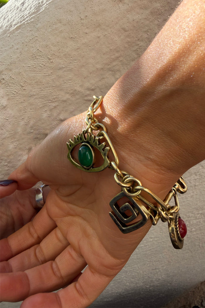 Brushed gold Deloris Charm Bracelet with chunky links and symbolic eye, spiral, and red stone charms on a person's wrist.