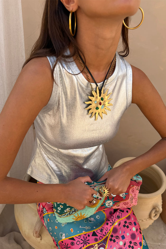 Iconic gold-tone Deloris Sun Necklace with semi-precious stones on a leather cord, styled over a silver metallic top.