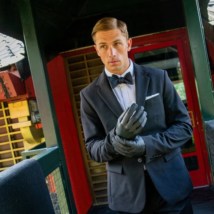 Man in formal attire with bow tie and gloves standing in front of a vintage vehicle.