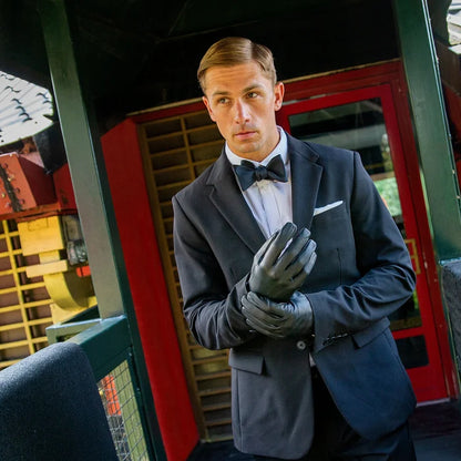 Man in formal attire with bow tie and gloves standing in front of a vintage vehicle.
