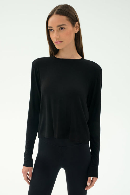 A woman wears the SPLITS59 Djuna Long Sleeve in black, paired with black leggings and standing against a plain light background—ideal women's activewear for cool weather workouts.
