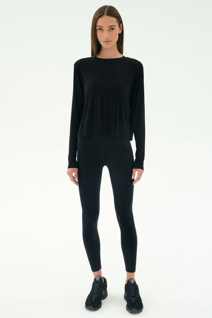 A person stands facing forward in SPLITS59's Djuna Long Sleeve - Black, paired with black leggings and sneakers—ideal activewear for cool weather workouts—against a plain white background.