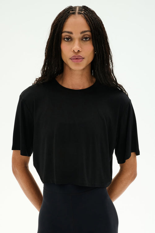 A person with long braided hair wears the SPLITS59 Djuna Oversized Crop Jersey Tee in black and black pants, standing against a plain white background.