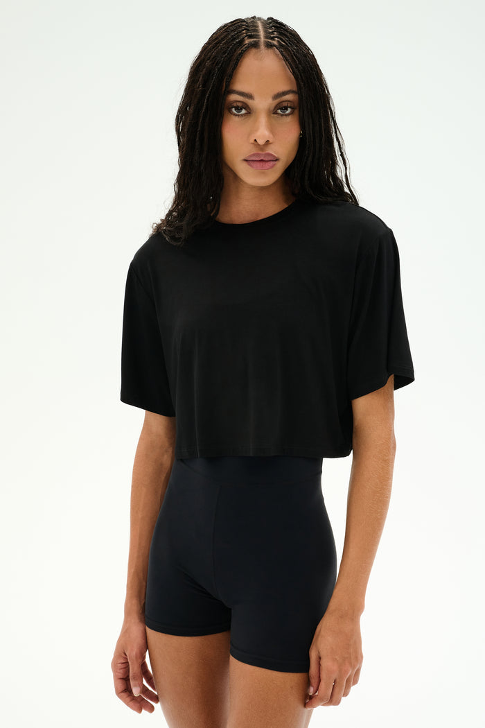 A person with long, braided hair wears the SPLITS59 Djuna Oversized Crop Jersey Tee in black and stretchy black high-waisted shorts, standing against a plain light background.