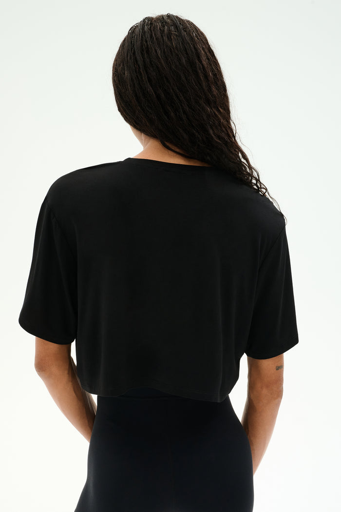 Person with long braided hair wears the SPLITS59 Djuna Oversized Crop Jersey Tee in black, a loose short-sleeve crop top shown from the back against a plain background.