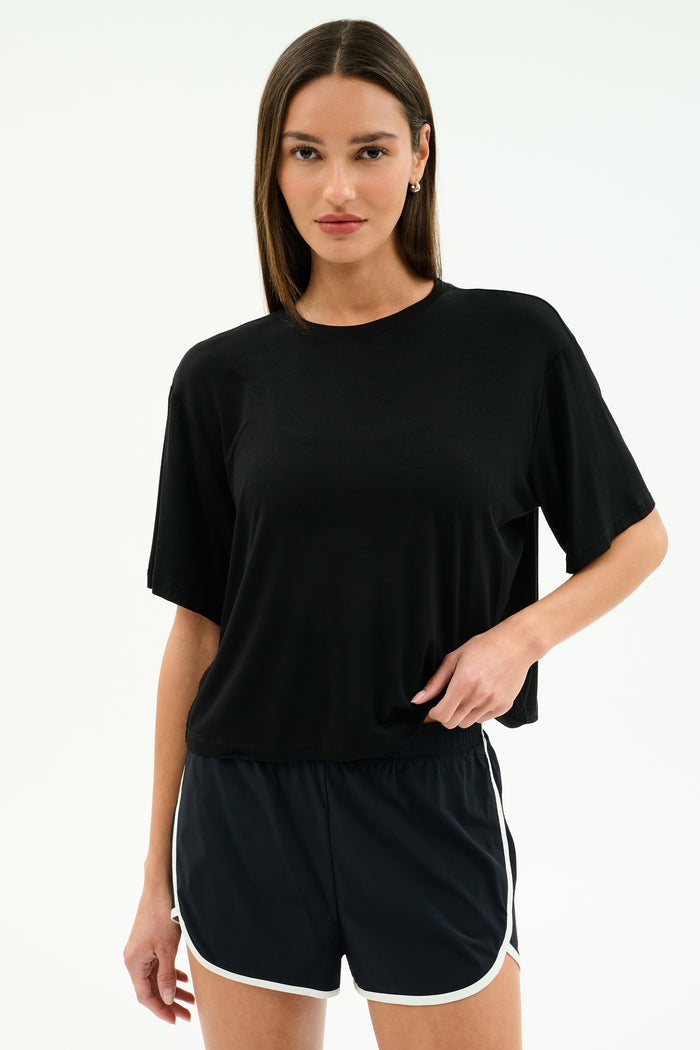 A woman stands against a plain background wearing the SPLITS59 Djuna Oversized Jersey Tee in black and black athletic shorts with white trim, perfect for gym workouts.