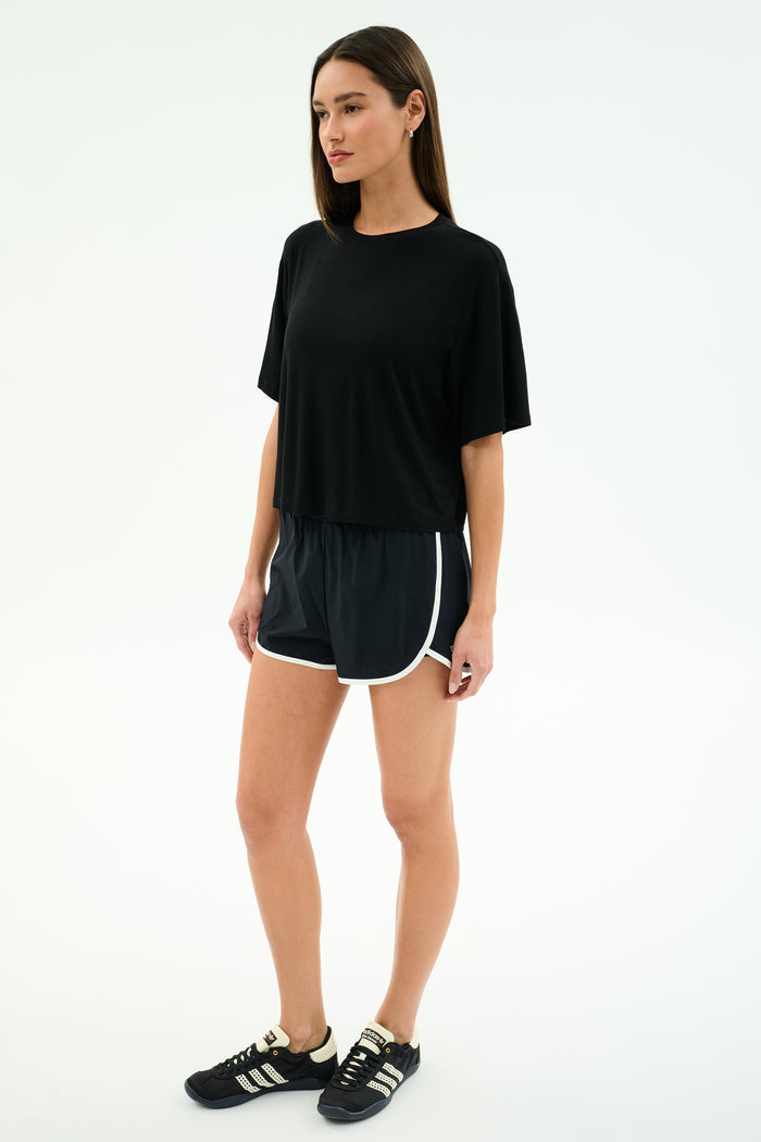 A woman stands against a plain background wearing the SPLITS59 Djuna Oversized Jersey Tee in black, black athletic shorts with white trim, and black Adidas sneakers—an effortless gym-ready look.