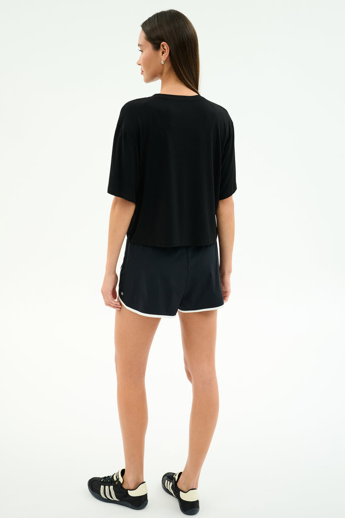 A person stands facing away, wearing the SPLITS59 Djuna Oversized Jersey Tee in black, paired with black shorts featuring white trim and black Adidas sneakers against a plain white background.