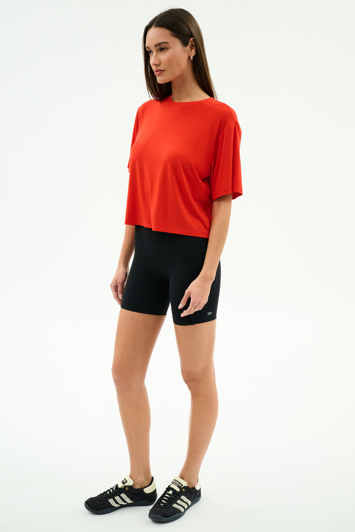 Woman standing against a plain white background, wearing the SPLITS59 Djuna Oversized Jersey Tee in Lotus Red, black bike shorts, and black Adidas sneakers—ideal for gym or outdoor workouts.