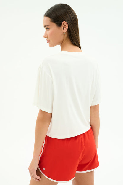A woman with straight brown hair stands facing away from the camera against a plain background, wearing the SPLITS59 Djuna Oversized Jersey Tee in white, paired with red athletic shorts—perfect for gym or outdoor workouts.