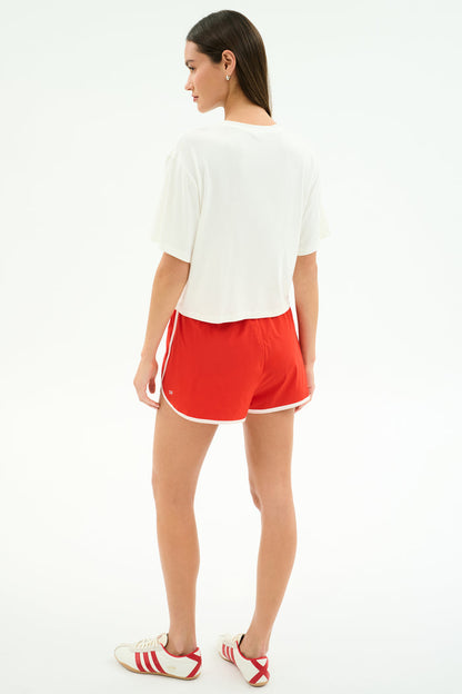 A woman stands facing away in the SPLITS59 Djuna Oversized Jersey Tee - White, paired with red athletic shorts with white trim and white sneakers with red stripes—an ideal outfit for gym workouts—set against a plain white background.
