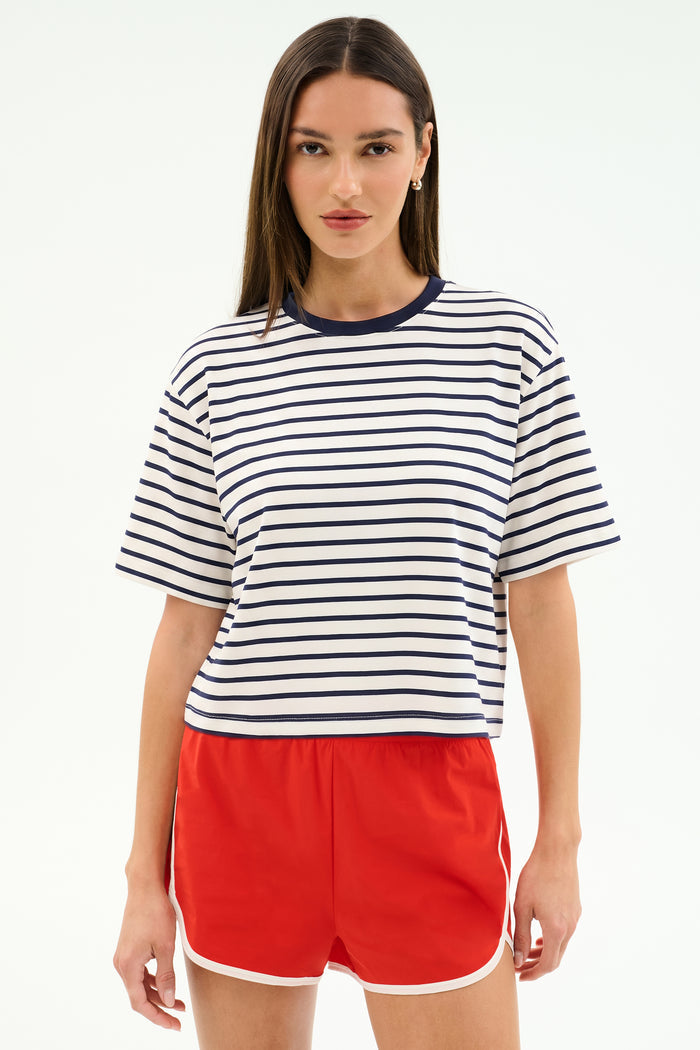 A woman stands facing forward, wearing the SPLITS59 Djuna Oversized Jersey Tee in White/Indigo Stripe with bright red athletic shorts.