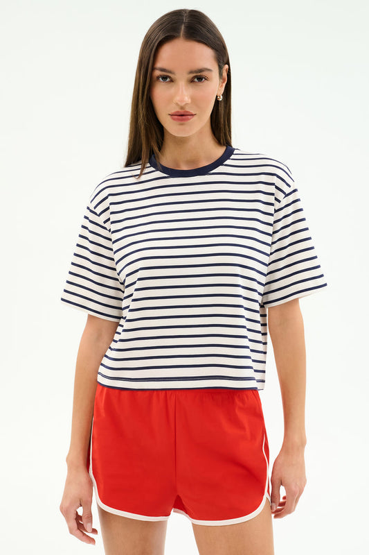 A woman stands facing forward, wearing the SPLITS59 Djuna Oversized Jersey Tee in White/Indigo Stripe with bright red athletic shorts.