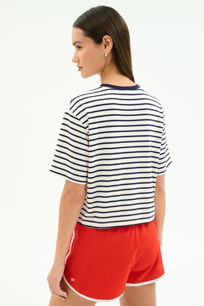 A woman stands with her back to the camera, wearing the SPLITS59 Djuna Oversized Jersey Tee in White/Indigo Stripe and red athletic shorts, ideal for gym workouts, set against a plain light background.