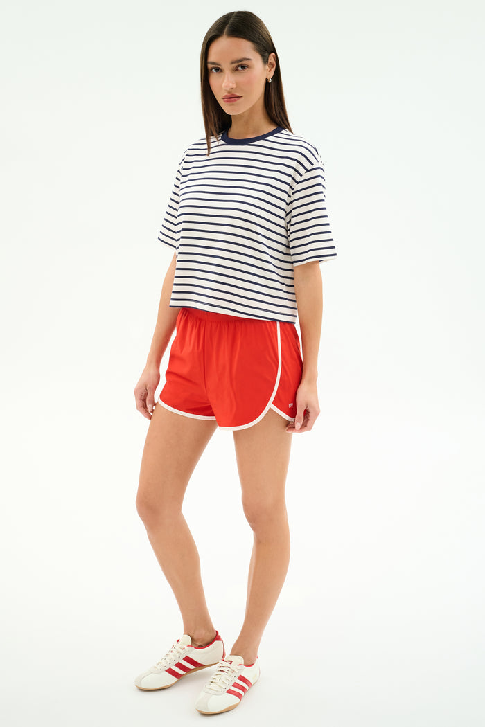 A woman stands facing forward in a SPLITS59 Djuna Oversized Jersey Tee in white/indigo stripe, red athletic shorts with white trim, and white sneakers with red stripes—perfect for gym or outdoor workouts.
