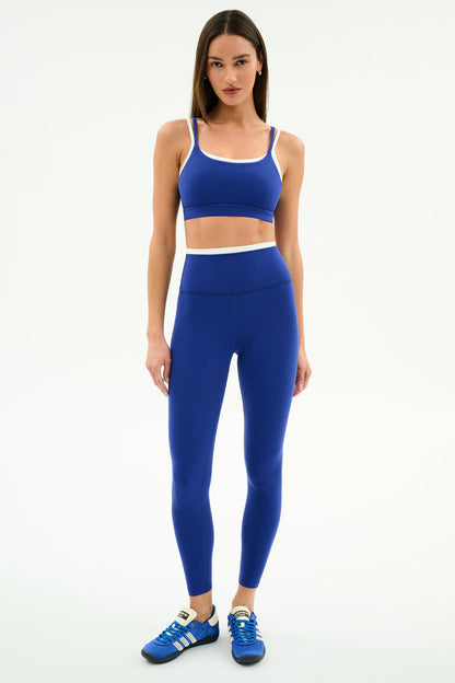 A woman faces forward in the SPLITS59 Dual High Waist Airweight 7/8 leggings in Cobalt/White with a matching sports bra and blue Adidas sneakers—perfect for hot yoga—against a plain white background.