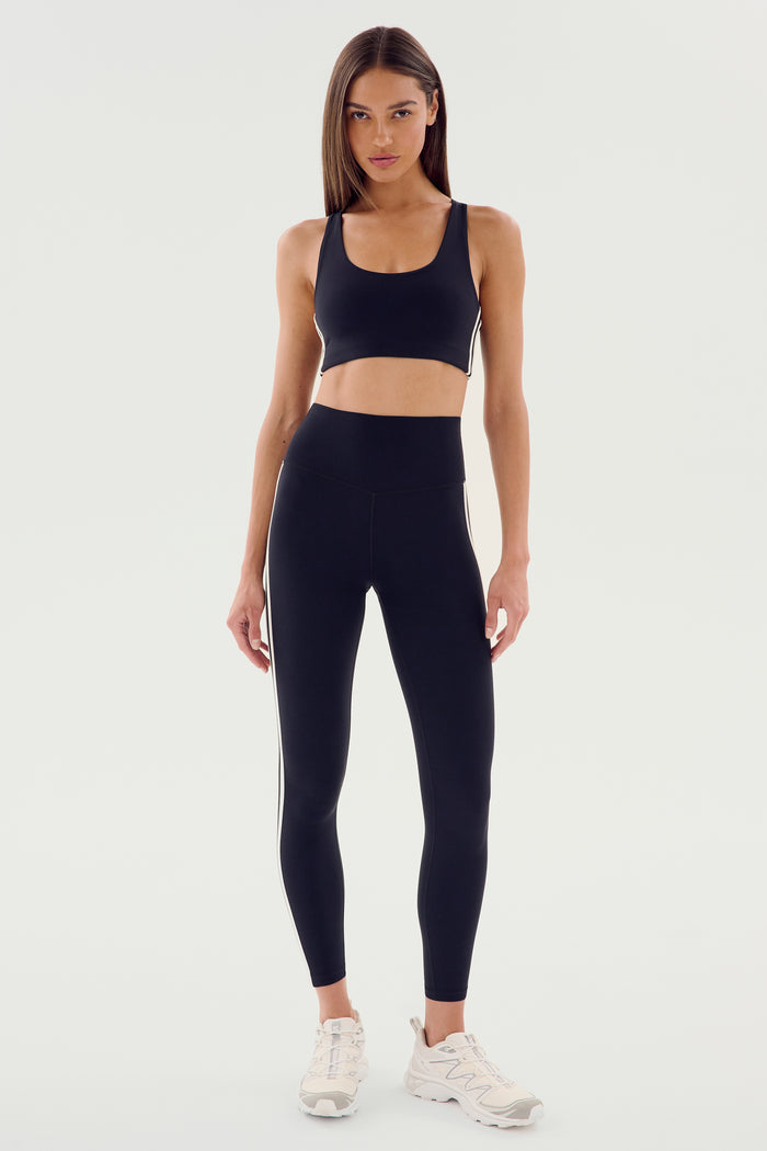 A person standing and wearing the SPLITS59 Ella Airweight Bra in Black/White paired with matching leggings featuring white side stripes and white sneakers, set against a plain background.