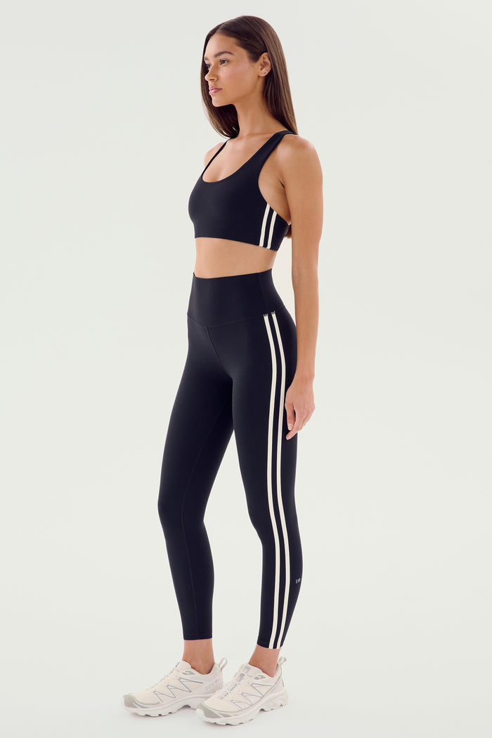 A woman wearing the SPLITS59 Ella Airweight Bra in black and white, along with black leggings featuring white stripes, stands in a side profile against a plain background. She completes her look with white sneakers.