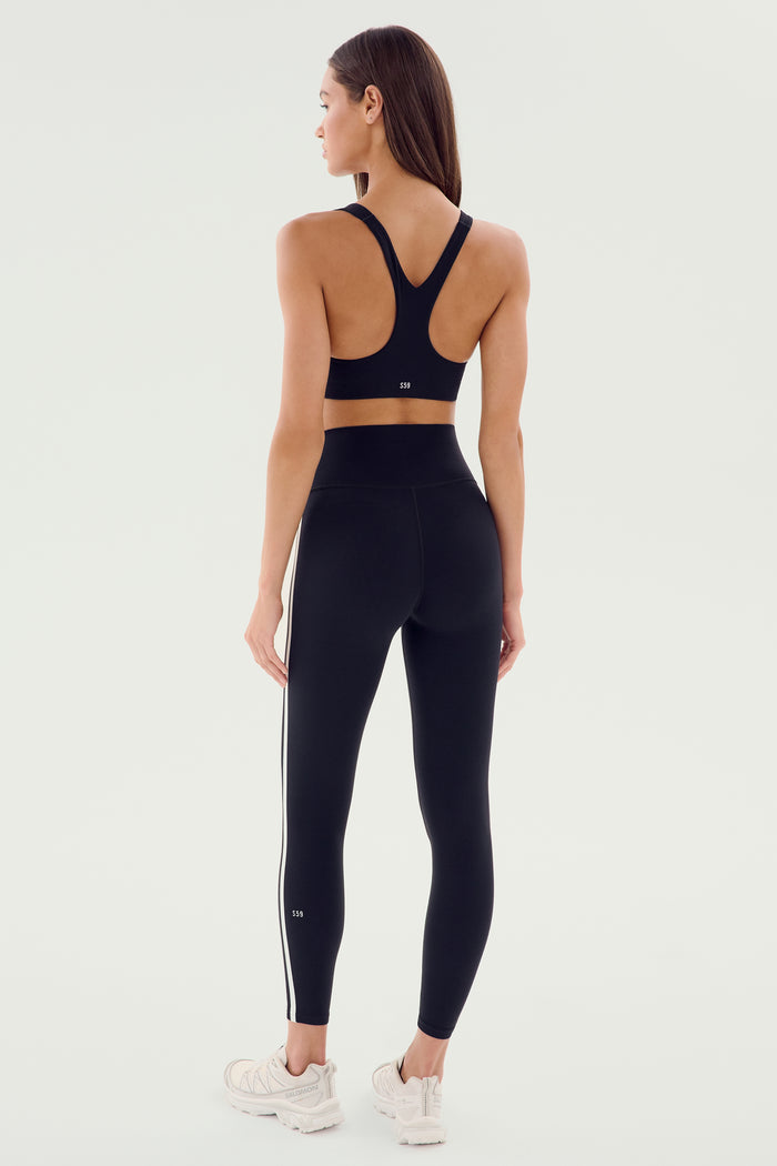 A person stands facing away, wearing the SPLITS59 Ella Airweight Bra in Black/White, high-waisted black leggings with white stripes, and white athletic shoes, ready for a hot yoga session.