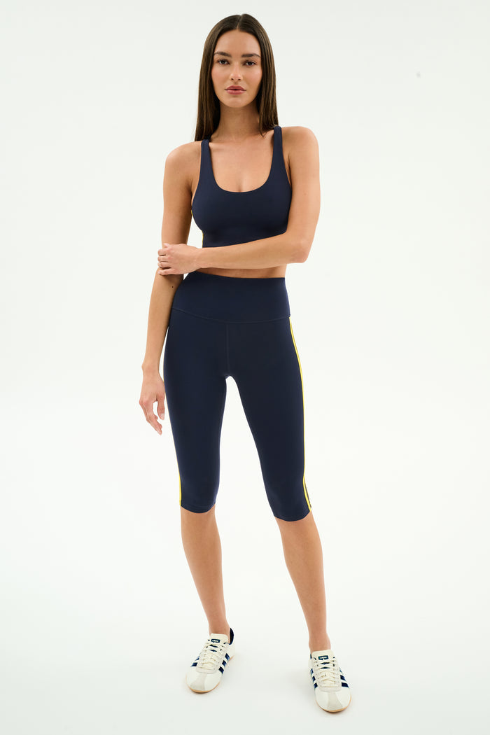 A woman stands facing forward in the SPLITS59 Ella Airweight Bra in Indigo/Bright Yellow, paired with matching capri leggings and white sneakers, all against a plain white background.
