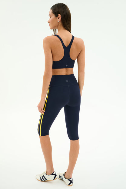 A woman stands facing away, wearing the SPLITS59 Ella Airweight Bra in Indigo/Bright Yellow and matching capri leggings, paired with white sneakers with black stripes, set against a plain background for an activewear look.