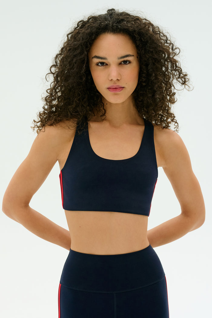 A woman with curly hair stands against a plain background, wearing the SPLITS59 Ella Airweight Bra in Indigo/Pirate Red, paired with matching leggings. The set features red side stripes and is crafted from stretchy nylon-spandex fabric.