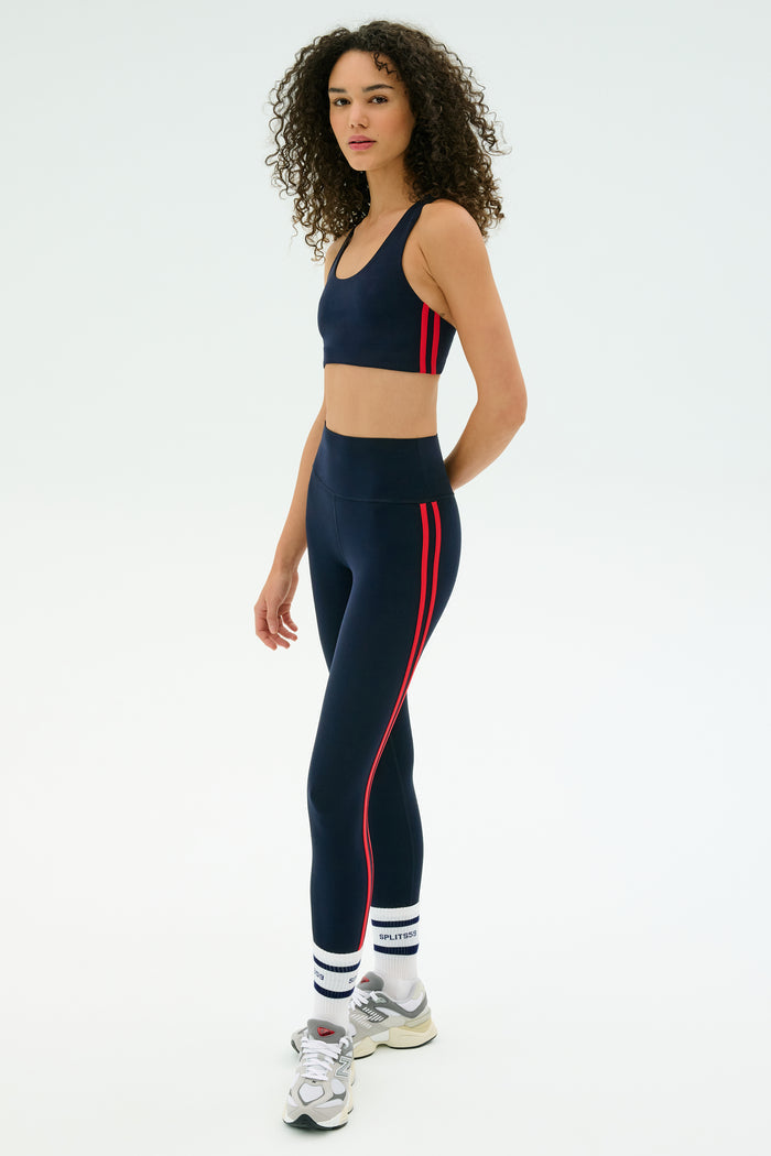 A person wears the SPLITS59 Ella Airweight Bra in Indigo/Pirate Red, paired with matching leggings, white socks, and silver sneakers, standing against a plain light background.