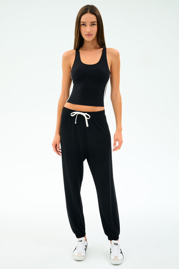 A woman faces forward in the SPLITS59 Ella Airweight Tank - Black/White, paired with black jogger pants and white sneakers, set against a plain white background.