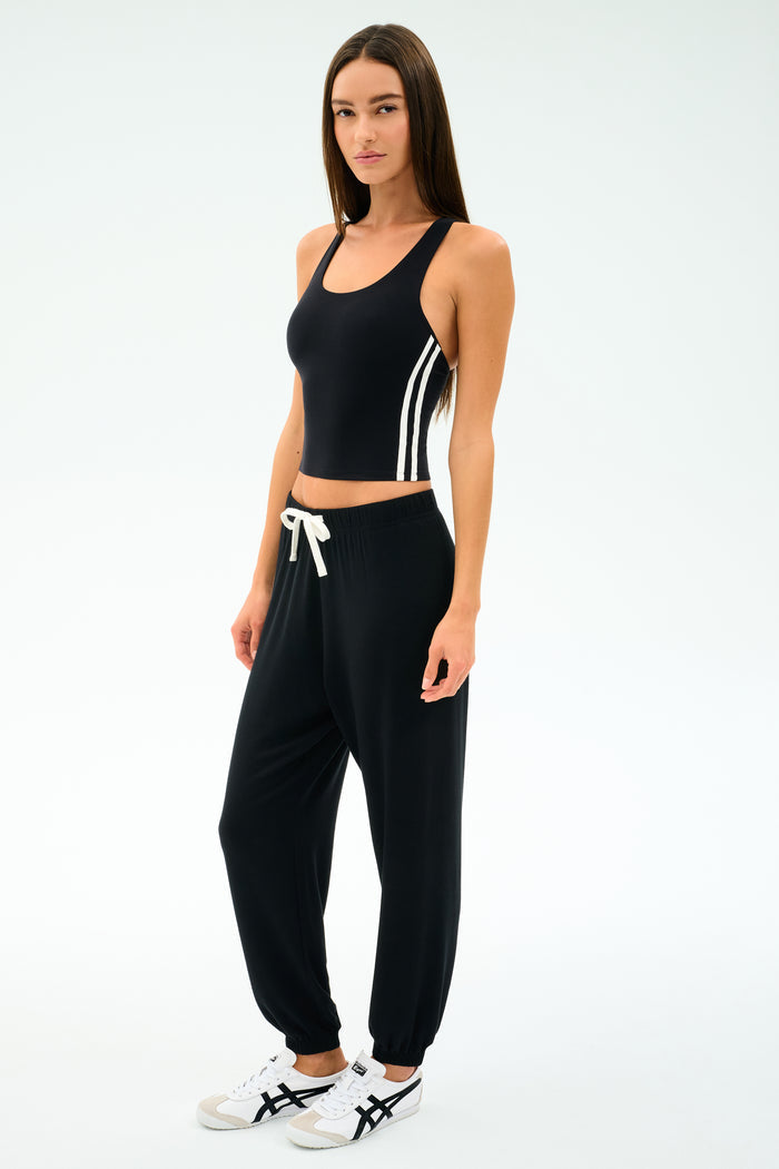 A woman faces forward on a white background, wearing the SPLITS59 Ella Airweight Tank in Black/White with side stripes, black nylon-spandex jogger pants, and white sneakers.
