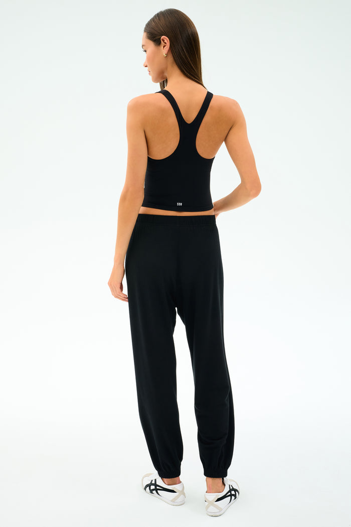 A woman faces away wearing the SPLITS59 Ella Airweight Tank in Black/White and jogger pants made from a soft nylon-spandex blend, paired with white sneakers featuring black accents.