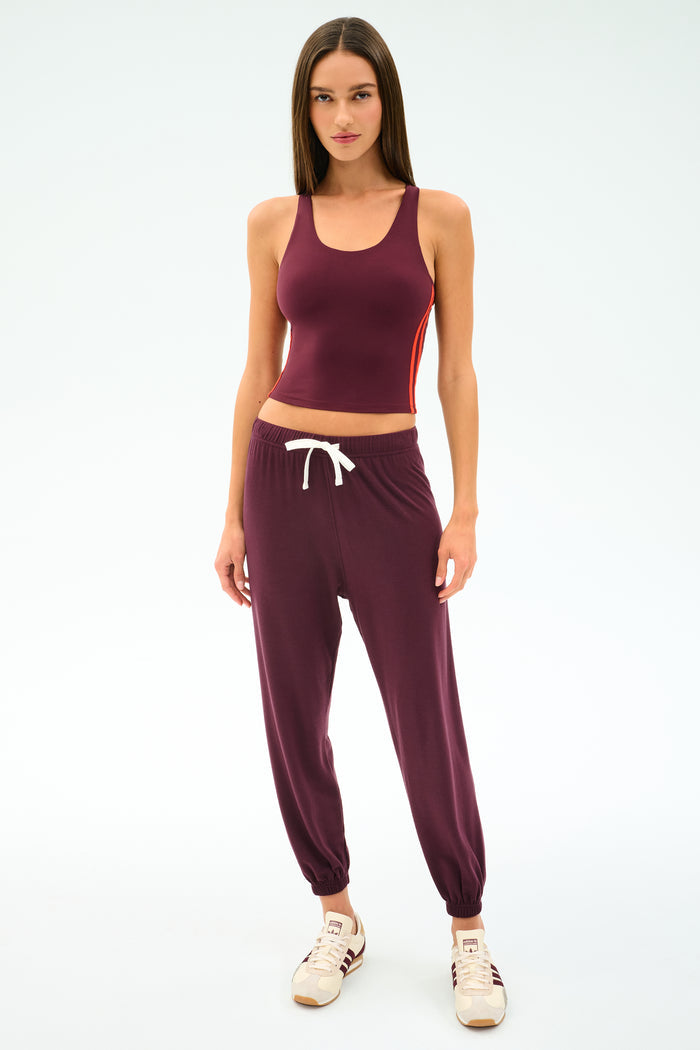 A woman faces forward in the SPLITS59 Ella Airweight Tank in Dark Cherry/Valencia, paired with matching joggers featuring a white drawstring and beige sneakers, all set against a plain white background.