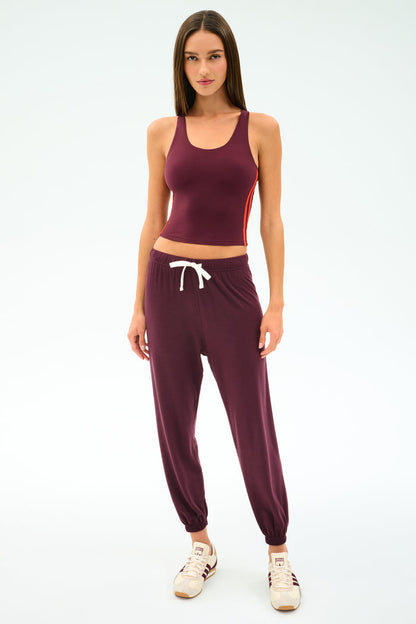 A woman faces forward in the SPLITS59 Ella Airweight Tank in Dark Cherry/Valencia, paired with matching joggers featuring a white drawstring and beige sneakers, all set against a plain white background.