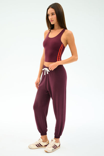 A woman wears the SPLITS59 Ella Airweight Tank in Dark Cherry/Valencia with matching jogger pants and white sneakers, standing against a plain light background.