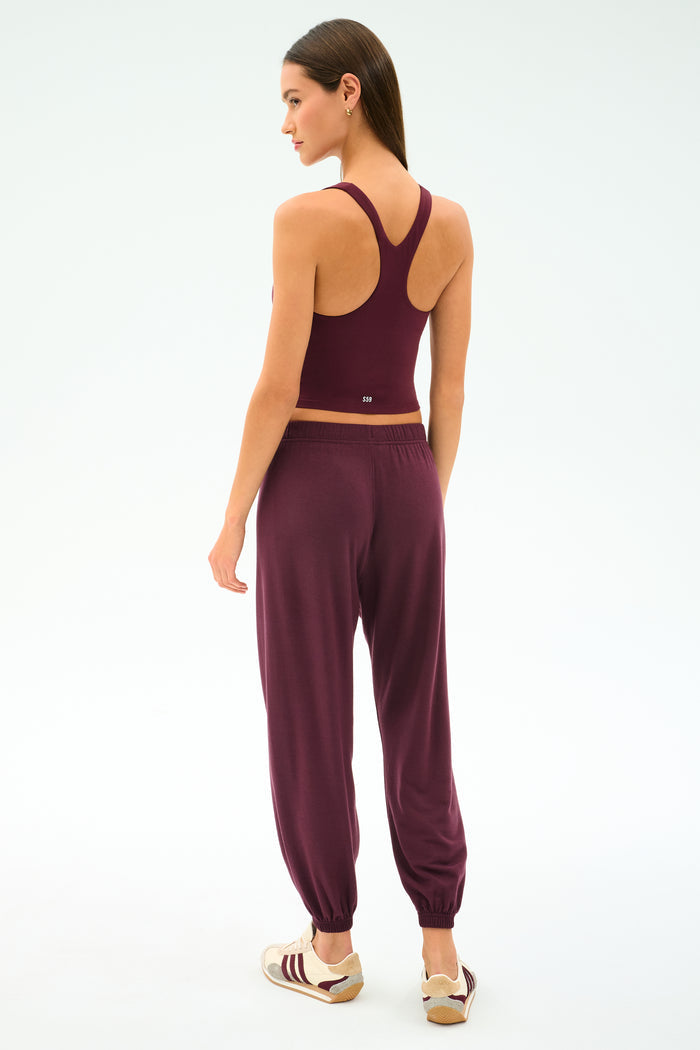 A woman faces away, wearing the SPLITS59 Ella Airweight Tank in Dark Cherry/Valencia, made from stretchy nylon spandex, paired with jogger pants and sneakers against a plain white background.