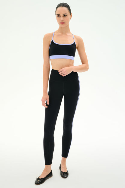 A woman stands against a plain background wearing SPLITS59's Ella High Waist Airweight 7/8 leggings in Black/Purple Haze, paired with a black sports bra and black ballet flats.