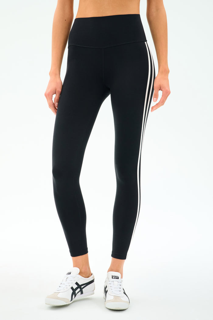 Someone in SPLITS59 Ella High Waist Airweight 7/8 leggings in Black/White with side stripes and white sneakers stands against a light background—ideal for hot yoga or everyday wear.