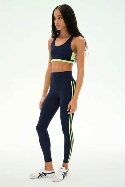 A woman poses against a plain light background wearing the SPLITS59 Ella High Waist Airweight 7/8 leggings and matching sports bra in Indigo/Limeade, styled with white sneakers.