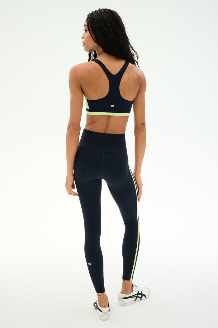 A person stands facing away against a plain white background, wearing the SPLITS59 Ella High Waist Airweight 7/8 leggings in Indigo/Limeade, paired with a navy sports bra and white sneakers.