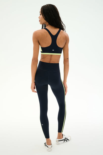 A person stands facing away against a plain white background, wearing the SPLITS59 Ella High Waist Airweight 7/8 leggings in Indigo/Limeade, paired with a navy sports bra and white sneakers.