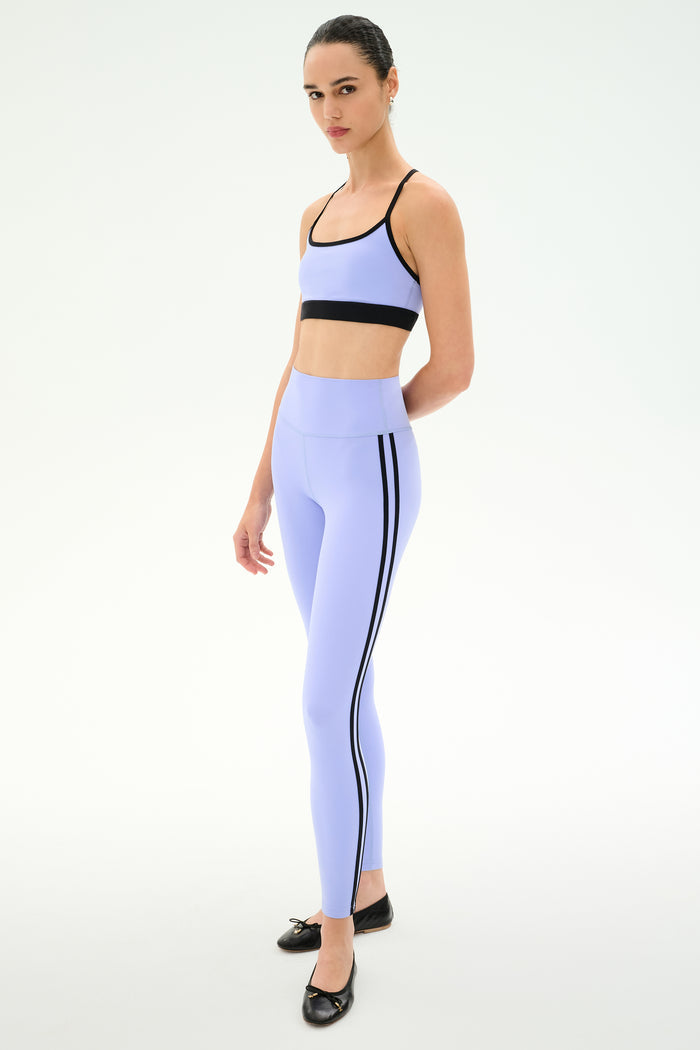 A person wears the SPLITS59 Ella High Waist Airweight 7/8 in Purple Haze/Black, paired with a matching light purple sports bra and black ballet flats, standing against a plain white background.