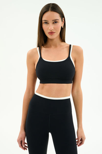 A woman wears the SPLITS59 Elliah Airweight Double Layer Bra in Black/White and high-waisted black nylon leggings, posing against a plain white background.
