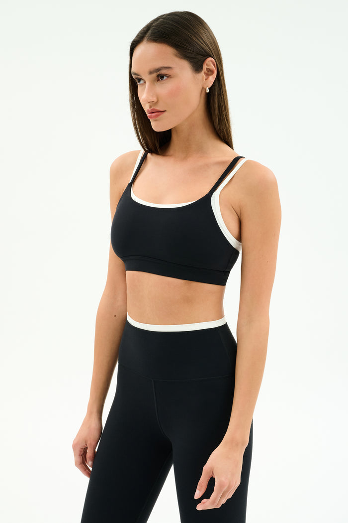 A woman wears the SPLITS59 Elliah Airweight Double Layer Bra in Black/White and high-waisted leggings, posing against a plain white background.