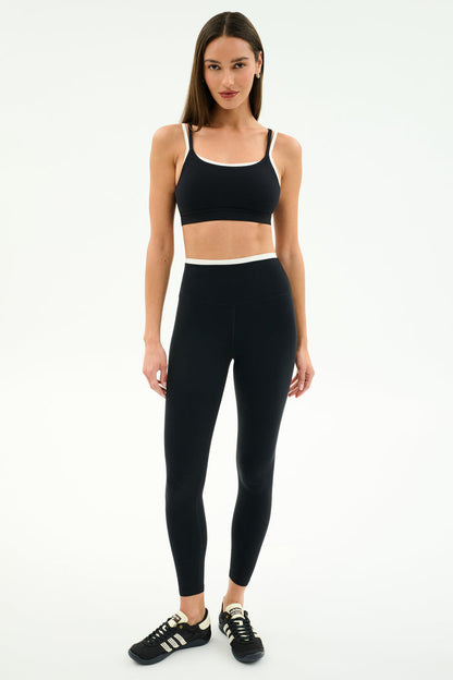 A woman stands against a plain white background wearing the SPLITS59 Elliah Airweight Double Layer Bra in Black/White, paired with matching high-waisted leggings and black athletic shoes.